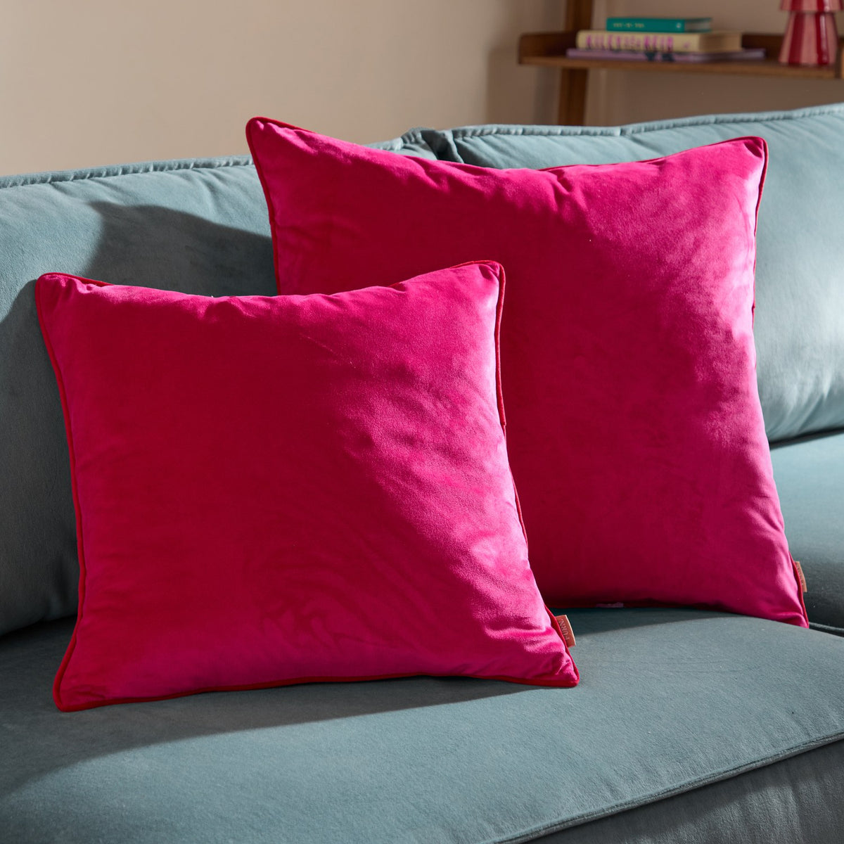Two Meridian square velvet cushions in hot pink with red piped trim, shown side by side on a light blue sofa in a living room setting.