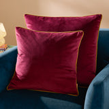 Two square Meridian velvet cushions in maroon with contrasting moss green piped edges, styled on a teal armchair against a neutral background.