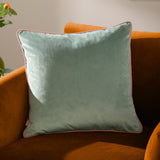 Meridian Plain Velvet Cushion in mineral blue with blush pink piped edging, shown square on an orange velvet sofa.