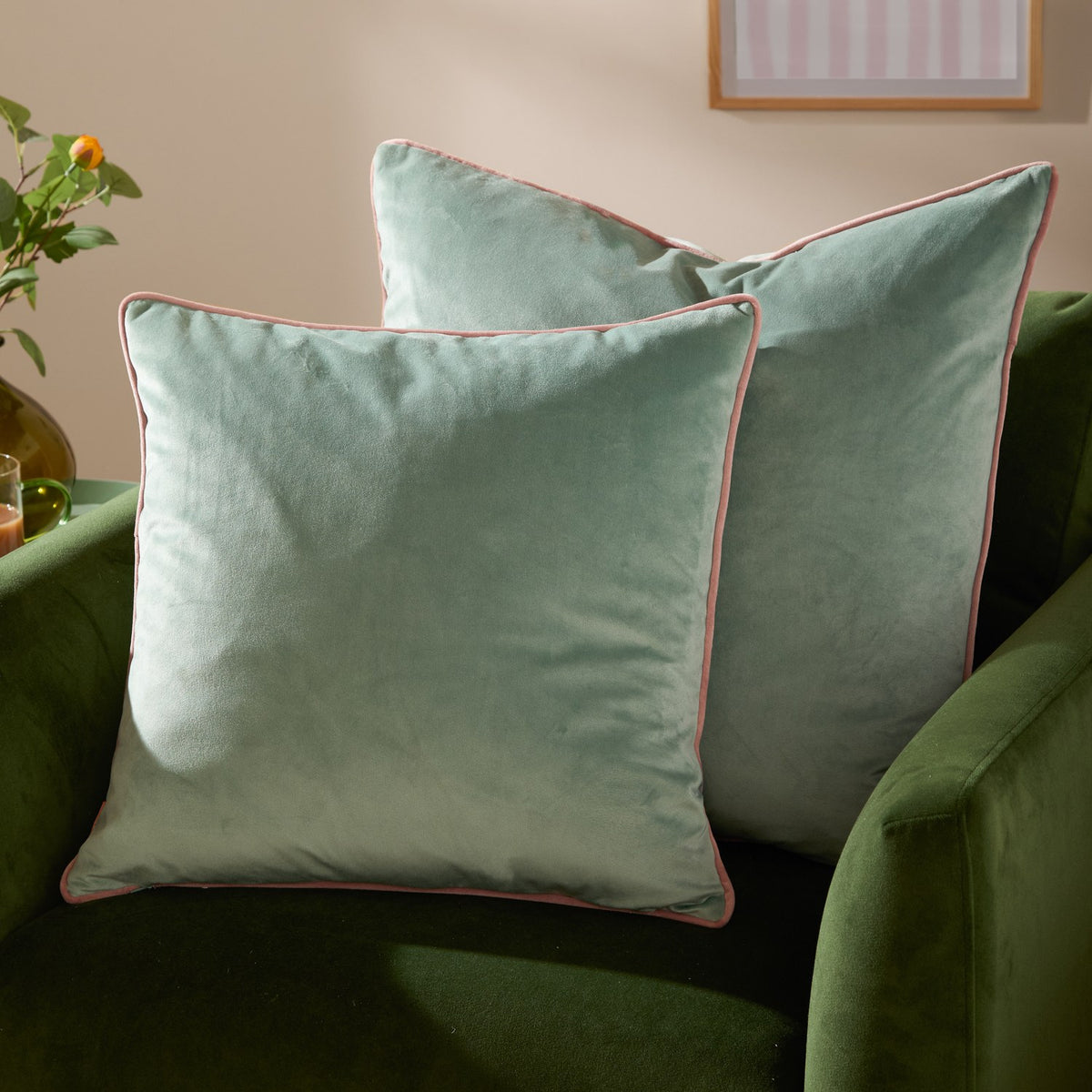 Two Meridian Plain Velvet Cushions in mineral blue with blush pink piped edges, styled on a green velvet armchair in a living room setting.