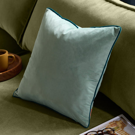 Meridian Plain Velvet Cushion in mineral blue with teal piped edge, square shape, shown on an olive green sofa beside a tray and mug.