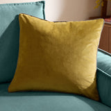 Meridian Plain Velvet Cushion in mustard-yellow velvet with charcoal piped edges, shown on a teal sofa in a living room setting.