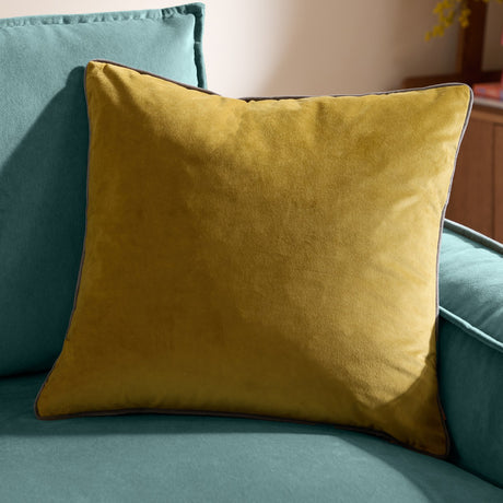 Meridian Plain Velvet Cushion in mustard-yellow velvet with charcoal piped edges, shown on a teal sofa in a living room setting.