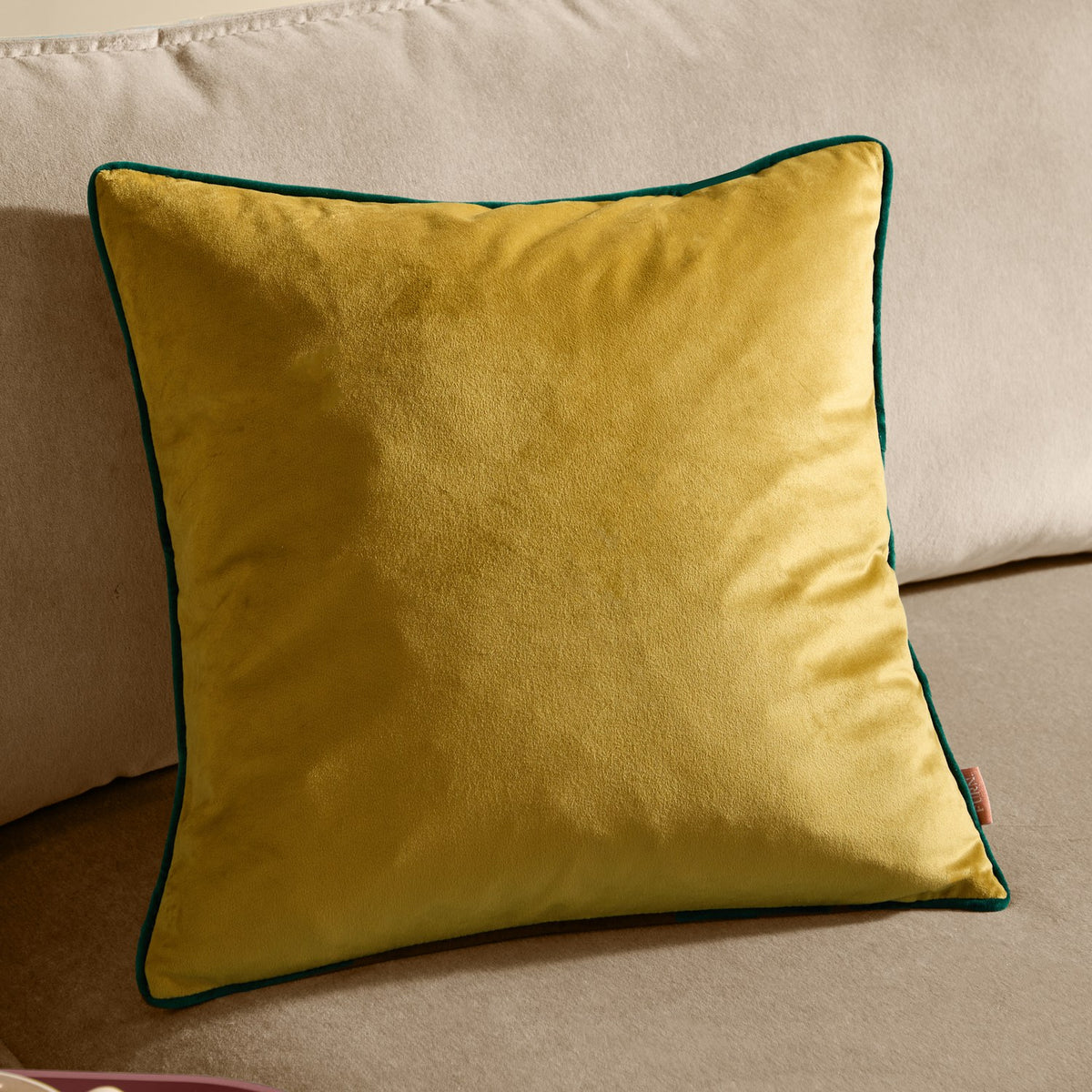 Meridian square velvet cushion in moss green with contrasting emerald piped edge, shown plain-fronted on a neutral beige sofa.