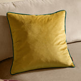 Meridian square velvet cushion in moss green with contrasting emerald piped edge, shown plain-fronted on a neutral beige sofa.
