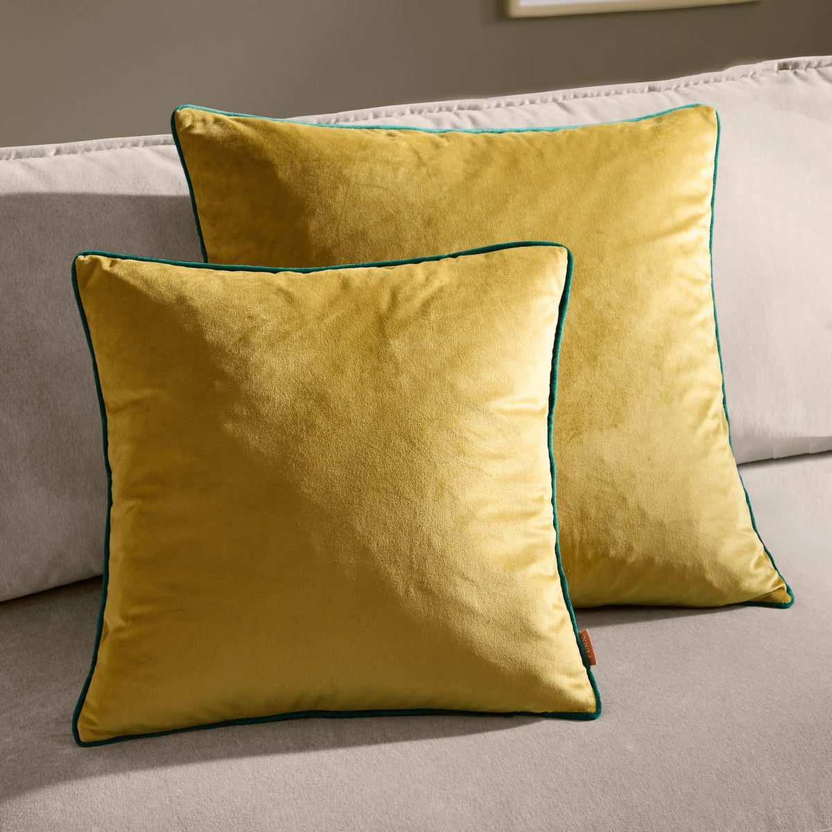 Two square Meridian velvet cushions in mustard-gold with contrasting emerald green piped edges, shown styled on a light grey sofa.