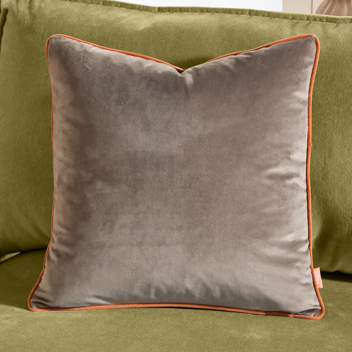 Meridian Plain Velvet Cushion in mocha brown with contrasting pumpkin orange piped edge, shown square-shaped on a green sofa.