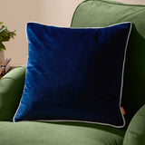 Meridian Plain Velvet Cushion in navy blue with silver-grey piped edging, shown on a green upholstered armchair in a living room setting.