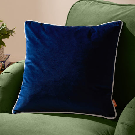 Meridian Plain Velvet Cushion in navy blue with silver-grey piped edging, shown on a green upholstered armchair in a living room setting.