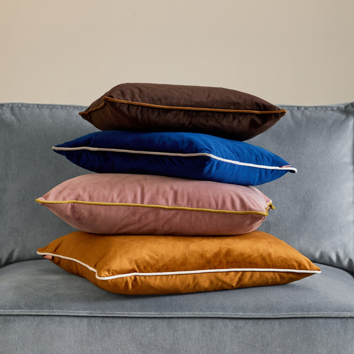 Meridian Plain Velvet Cushion in navy blue with silver-grey piped edge, shown velvet-textured and stacked with other cushions on a grey sofa.