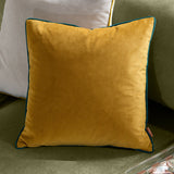 Meridian Plain Velvet Cushion in ochre with contrasting teal piped edging, shown on a green sofa against light cushions in a living room setting.