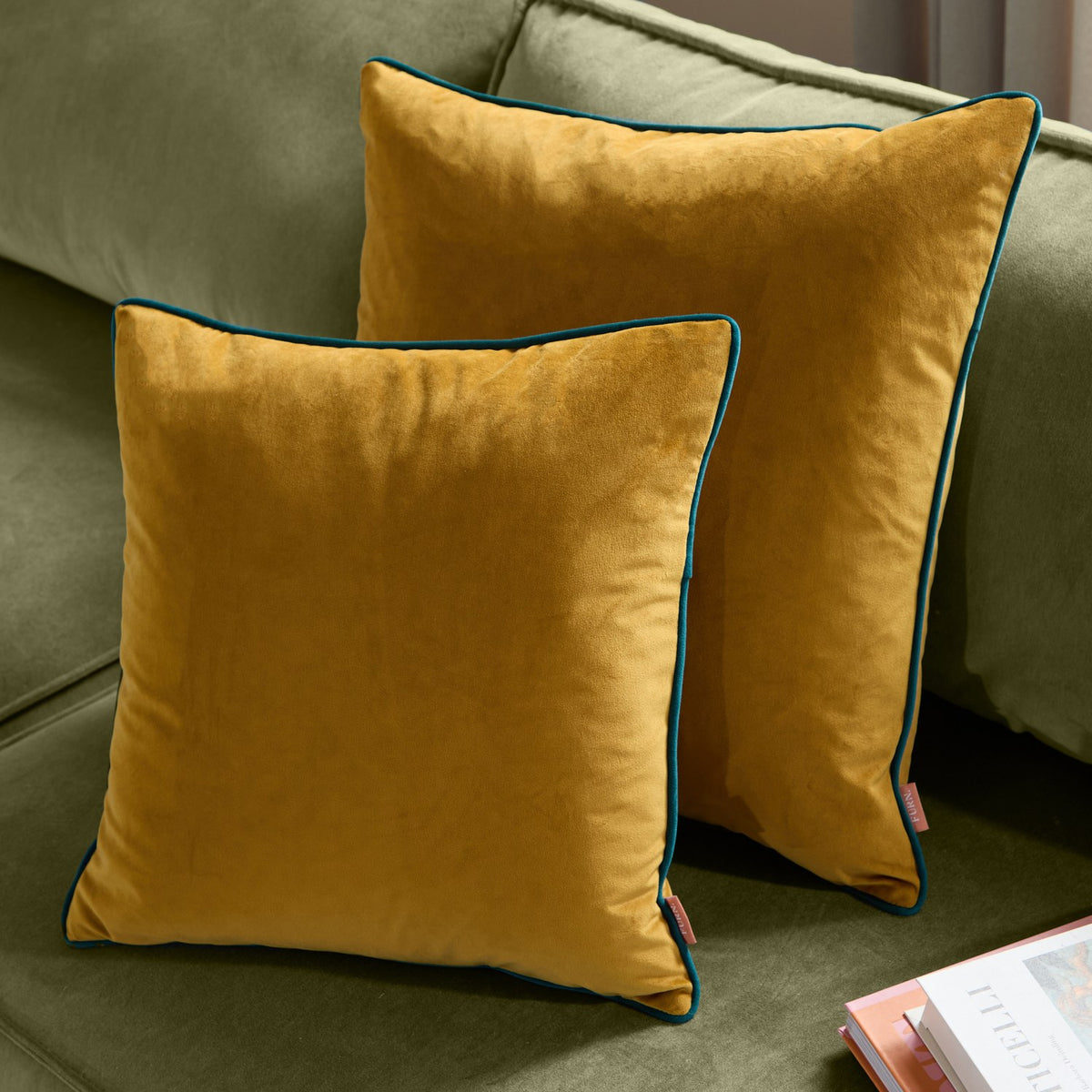 Meridian Plain Velvet Cushions in ochre with teal piped trim, shown in two square sizes on a green sofa alongside books.