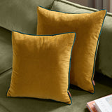 Meridian Plain Velvet Cushions in ochre with teal piped trim, shown in two square sizes on a green sofa alongside books.