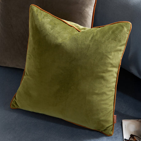 Meridian Plain Velvet Cushion in olive green with contrasting ginger piped trim, shown square-shaped on a blue sofa alongside a grey cushion.
