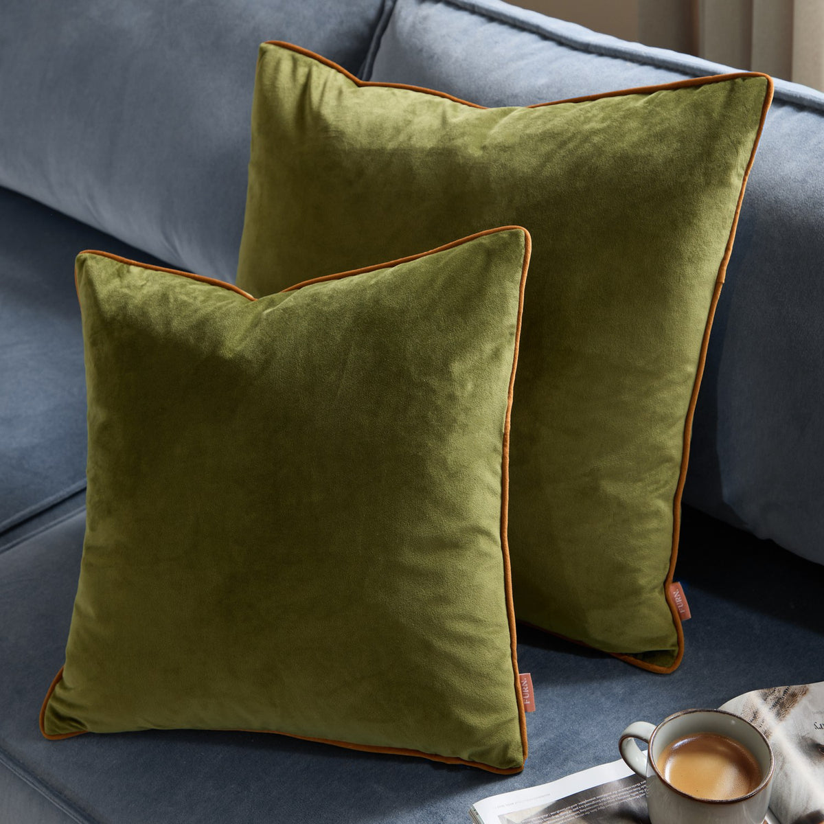 Two Meridian plain velvet cushions in olive green with contrasting ginger piping, shown on a blue sofa beside a mug and magazine.