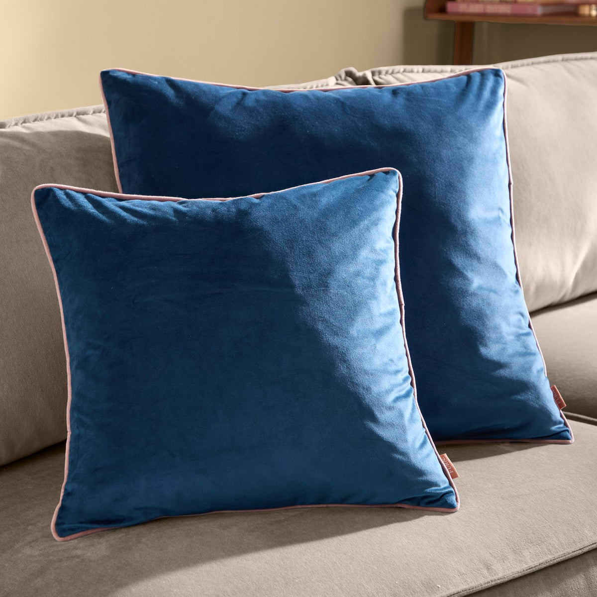 Two Meridian plain velvet square cushions in petrol blue with blush pink piped edges, styled on a grey sofa in a living room setting.