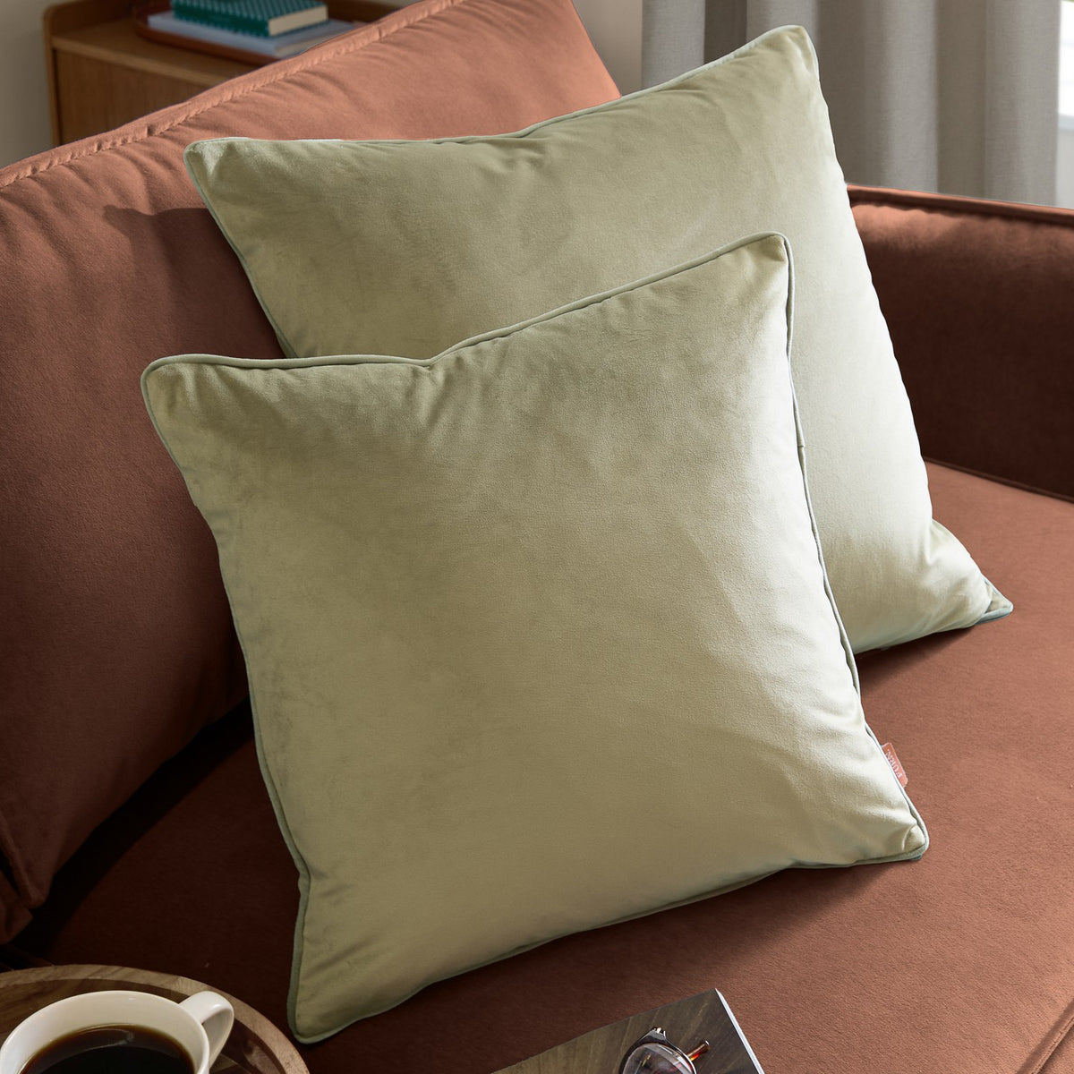 Two Meridian square velvet cushions in sage green with contrasting piped edges, styled on a rust-coloured sofa in a living room setting.