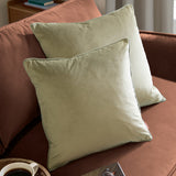Two Meridian square velvet cushions in sage green with contrasting piped edges, styled on a rust-coloured sofa in a living room setting.