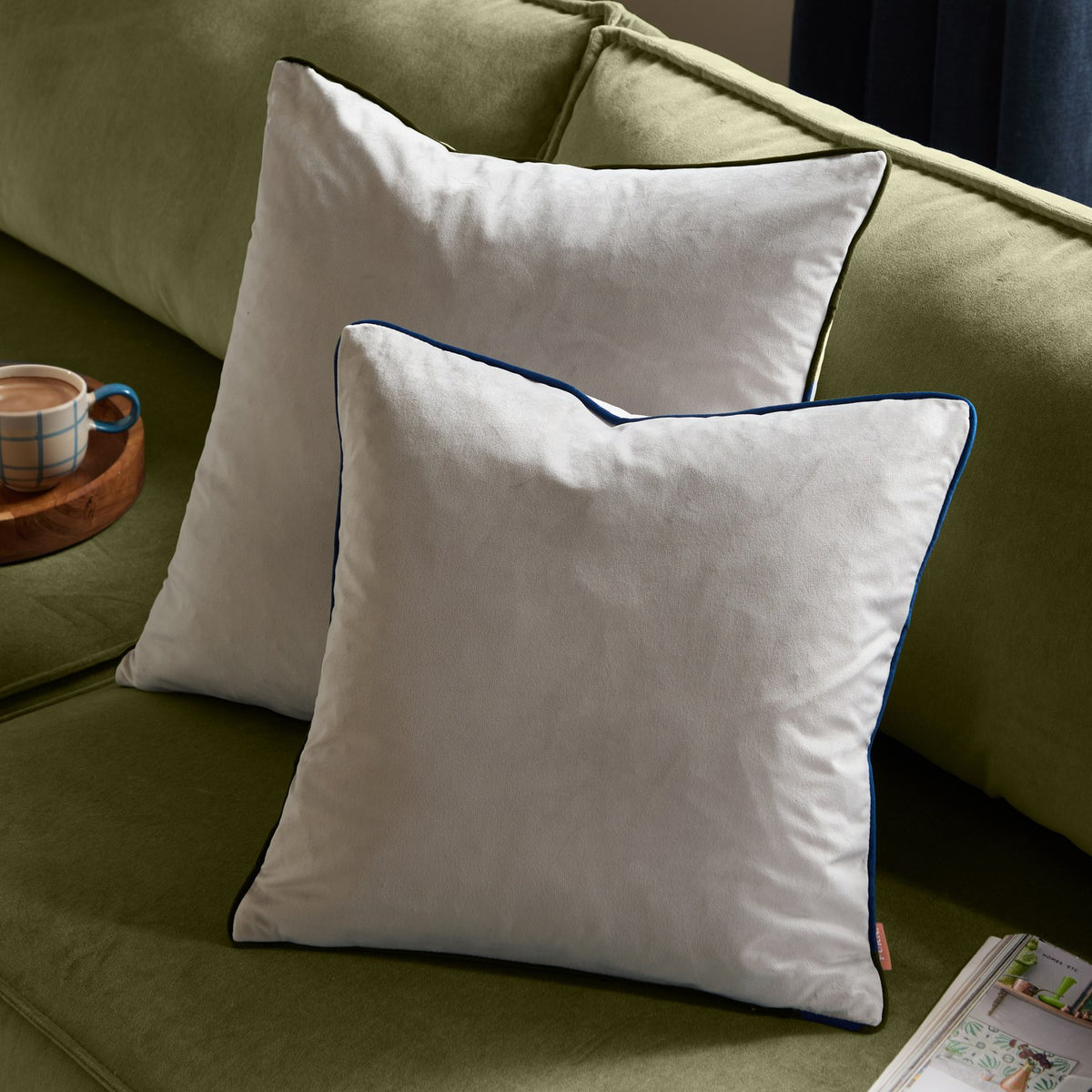 Two Meridian plain velvet cushions in silver grey with navy piped edges, styled on a green sofa beside a mug and tray.