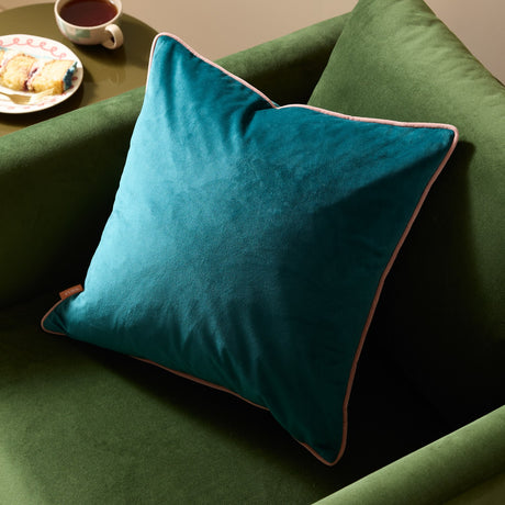 Meridian square cushion in deep teal velvet-feel fabric with contrasting blush pink piped edging, shown on a green velvet sofa.