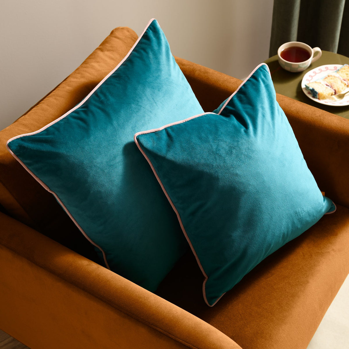 Two Meridian teal velvet square cushions with blush pink piped edging, styled on an orange velvet sofa in a living room setting.