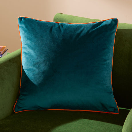 Meridian Plain Velvet Cushion in teal with clementine orange piped edging, shown on a green upholstered sofa in a living room setting.