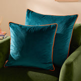 Two square Meridian velvet cushions in teal with clementine orange piped edges, styled on a green sofa in a living room setting.