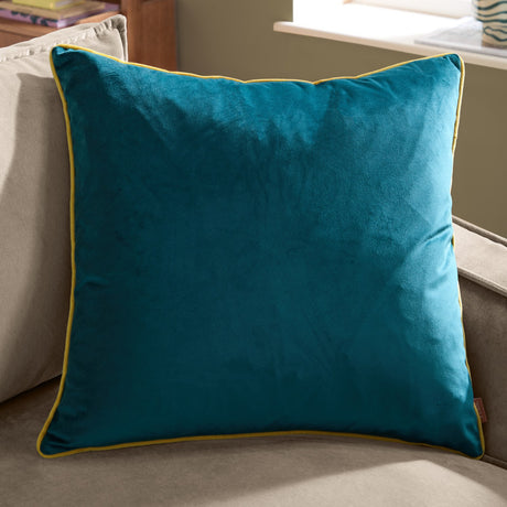 Meridian Plain Velvet Cushion in teal blue with contrasting yellow piped edge, shown square on a beige sofa in a living room setting.