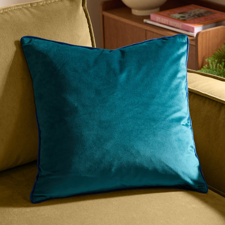 Meridian Plain Velvet Cushion in teal with contrasting navy piped trim, square shape, shown on a mustard sofa beside a wooden side table.