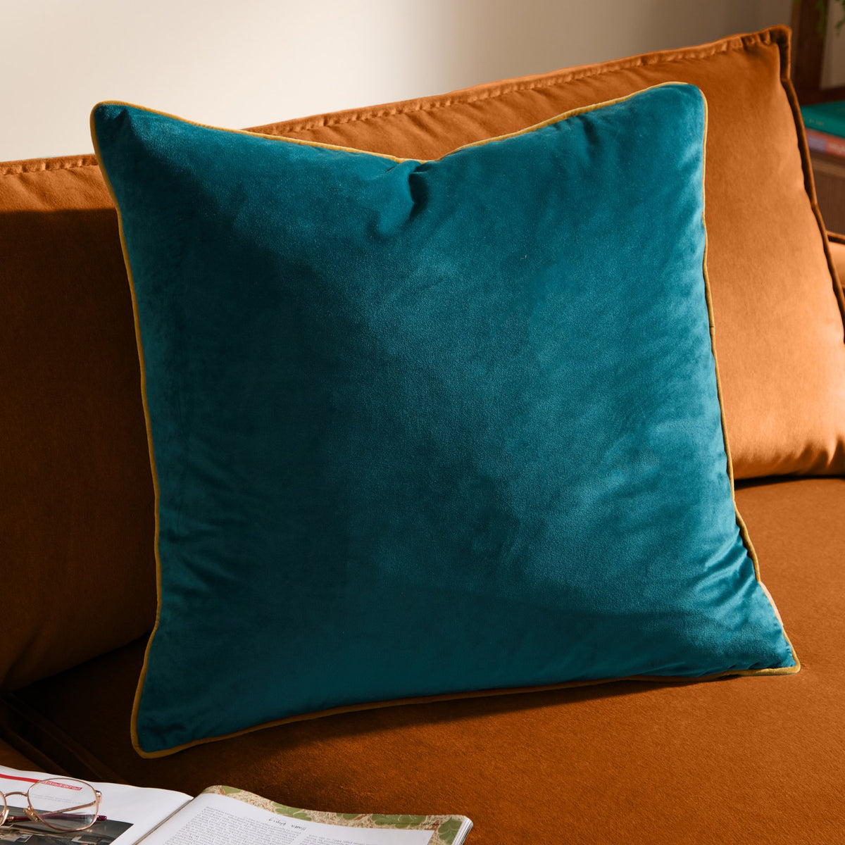 Meridian Plain Velvet Cushion in deep teal with ochre-gold piped trim, square shape, shown on an orange sofa beside an open book.