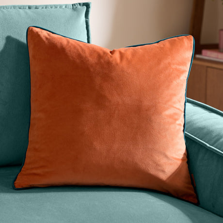 Meridian Plain Velvet Cushion in burnt orange with contrast teal piped edging, styled on a teal velvet sofa in a living room setting.