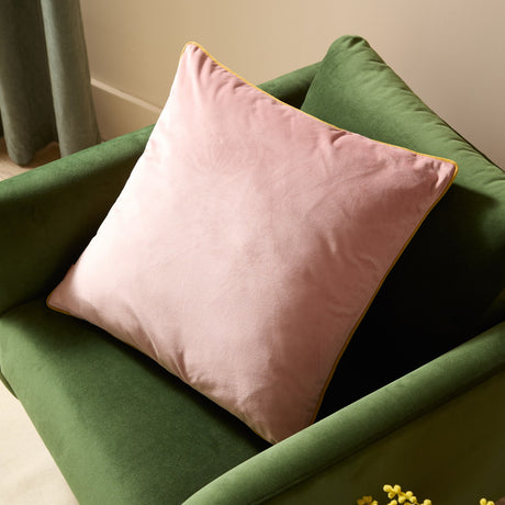Meridian Plain Velvet Cushion in dusty pink with gold piped edging, shown resting in the corner of a green velvet armchair.