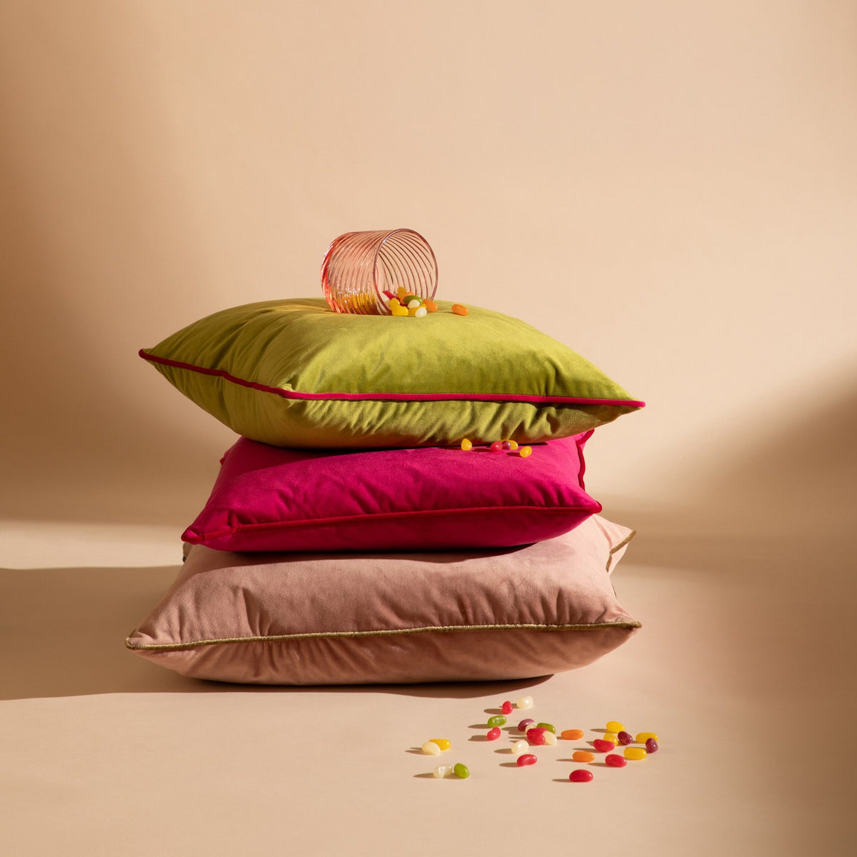 Meridian Plain Velvet Cushion in dusty pink with gold piped edging, shown at the base of a stacked pillow display with green and pink cushions and scattered jelly beans.