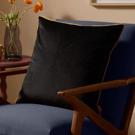 Meridian Plain Velvet Cushion in black with gold piping edging, square shape, shown on a navy armchair beside a wooden side table and vase of flowers.