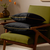 Two Meridian black velvet-feel cushions with gold piping edging, stacked on a green upholstered armchair with wooden frame in a living room setting.