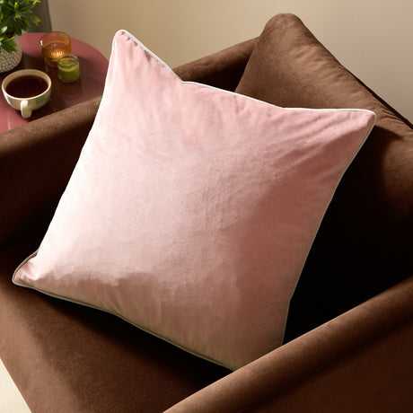 Meridian Plain Velvet Cushion in blush pink with contrasting light grey piped edging, square velvet-feel design shown on a brown upholstered chair.