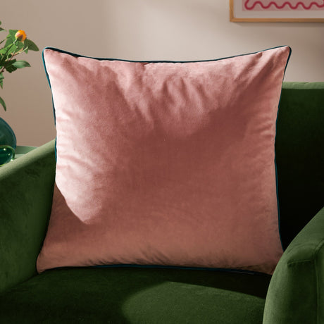 Meridian Plain Velvet Cushion in blush pink with contrasting teal piped edge, shown on a green velvet sofa in a living room setting.