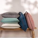 Meridian Plain Velvet Cushion in blush pink with contrasting teal piped edge, shown among grey, mint, white, and teal velvet cushions on a wooden bench.