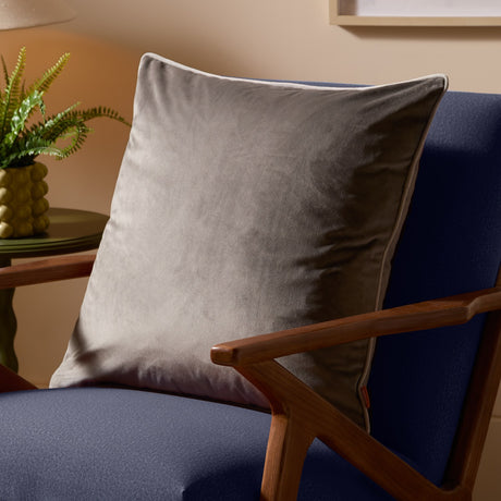 Meridian Plain Velvet Cushion in charcoal grey with dove silver piped edging, shown resting on a navy armchair in a living room setting.