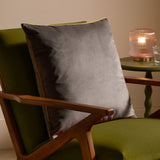 Meridian Plain Velvet Cushion in charcoal grey with moss green piped edging, styled on a green upholstered armchair with wooden arms beside a side table and lamp.