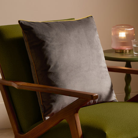 Meridian Plain Velvet Cushion in charcoal grey with moss green piped edging, styled on a green upholstered armchair with wooden arms beside a side table and lamp.
