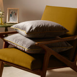 Two Meridian Plain Velvet Cushions in charcoal grey with contrasting yellow piped edges, stacked on a mustard armchair with wooden frame in a living room setting.