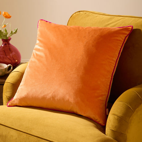 Meridian Plain Velvet Cushion in clementine orange with hot pink piped edging, shown on a mustard velvet armchair beside a vase of flowers.