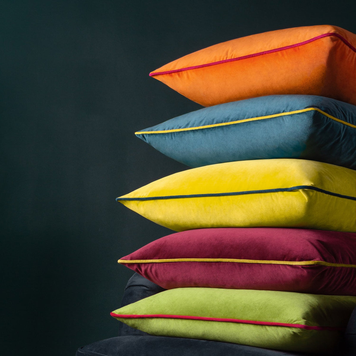 Meridian Plain Velvet Cushion in clementine orange with hot pink piped edging, shown top of a stack of velvet cushions in teal, yellow, and green against a dark background.