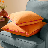 Two square Meridian velvet cushions in clementine orange with contrasting hot pink piped edges, stacked on a teal armchair beside a side table.