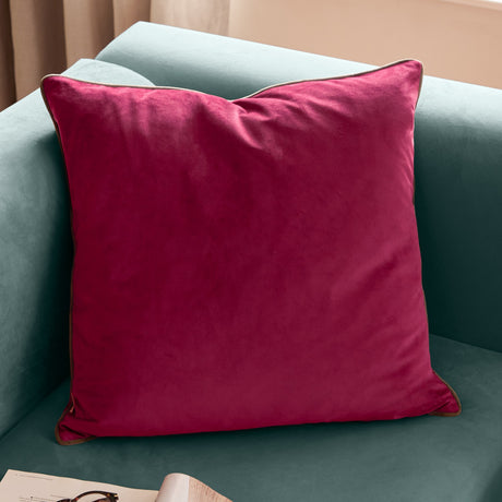 Meridian Plain Velvet Cushion in cranberry pink with a mocha piped edge, shown square-shaped on a teal sofa in a living room setting.