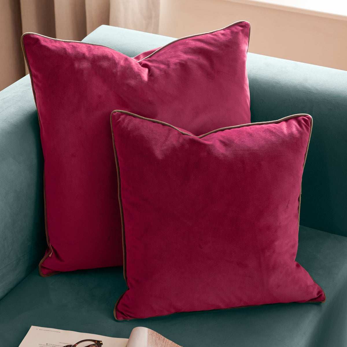 Two Meridian Plain Velvet Cushions in cranberry pink with mocha brown piped edges, styled on a teal velvet sofa.
