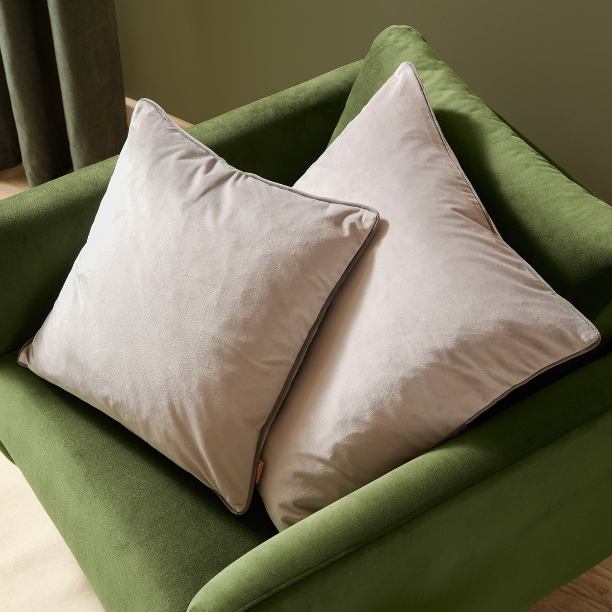 Two square Meridian velvet cushions in dove grey with charcoal piped edges, styled on a green velvet armchair.