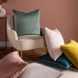 Meridian Plain Velvet Cushion in dove grey with charcoal piped edge, shown square on a cream armchair alongside pink, teal, and yellow velvet cushions in a living room setting.