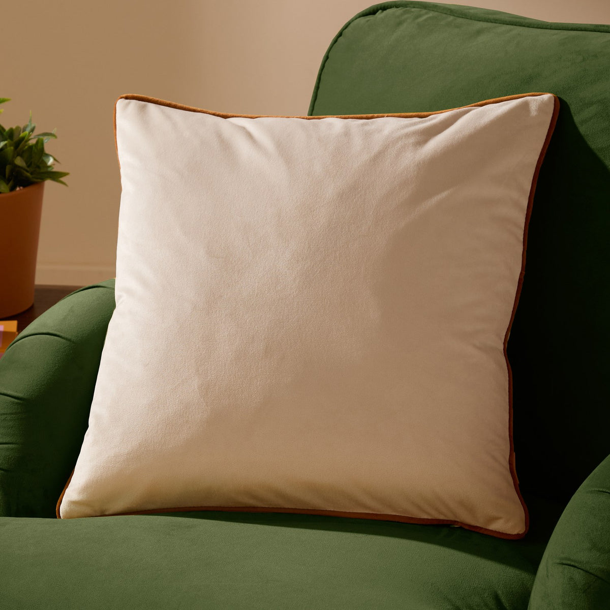 Meridian Plain Velvet Cushion in ecru with contrasting ginger piped edge, square scatter cushion shown on a green sofa.